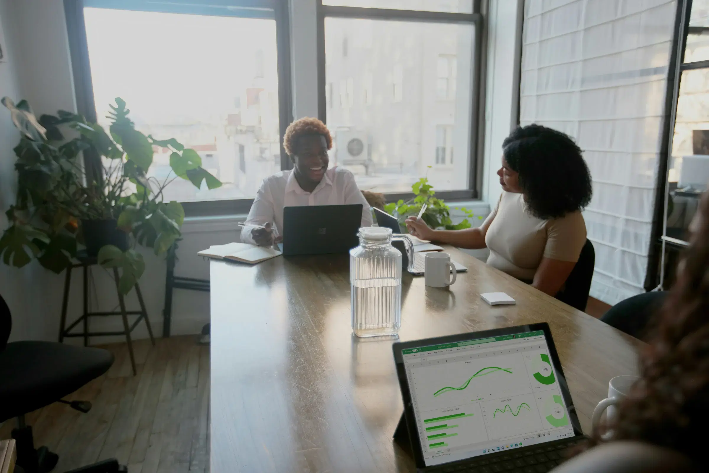 Two colleagues meeting at a long table by the window, a tablet showing a reports dashboard