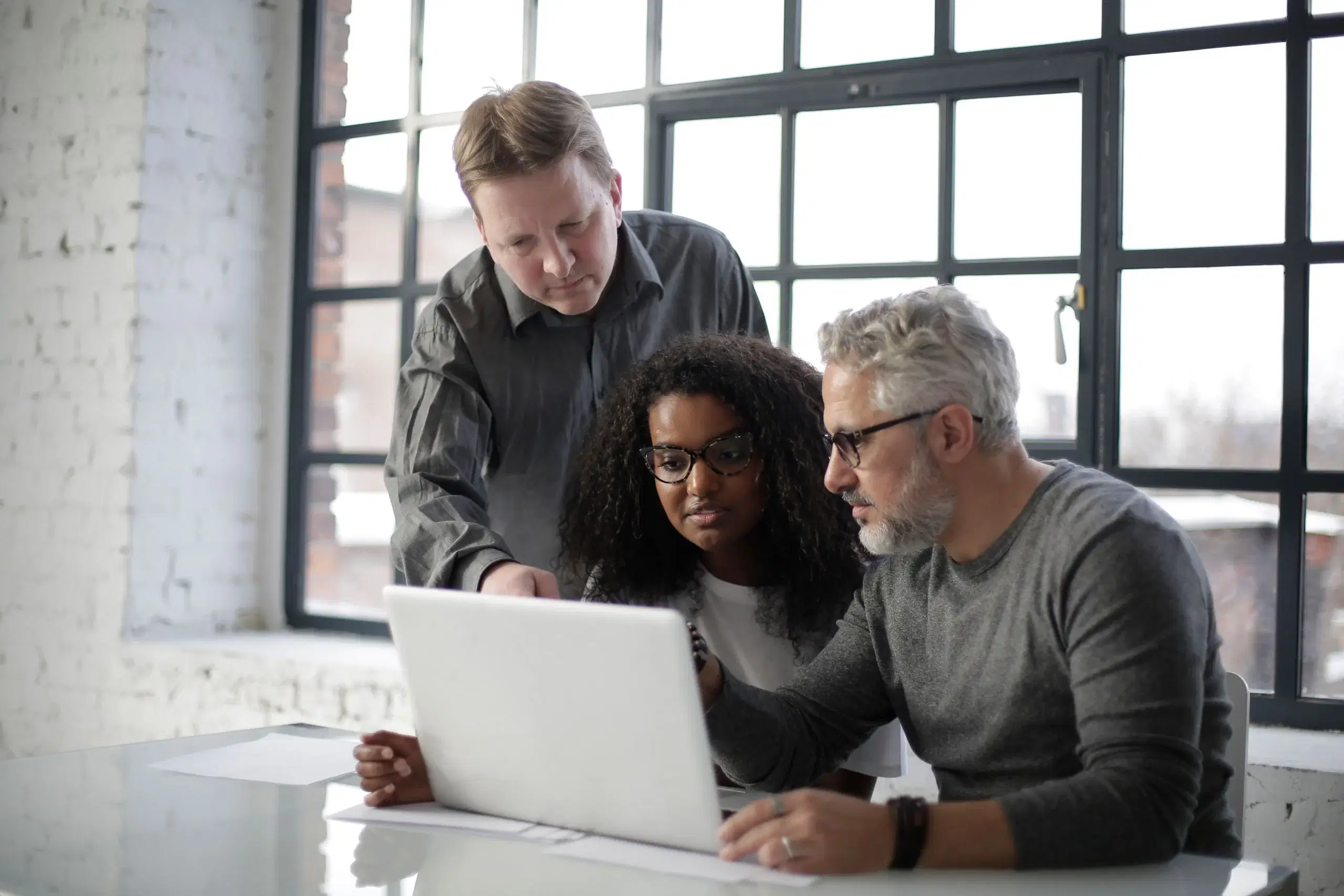 Three experienced colleagues working through something together on a laptop by a large office window